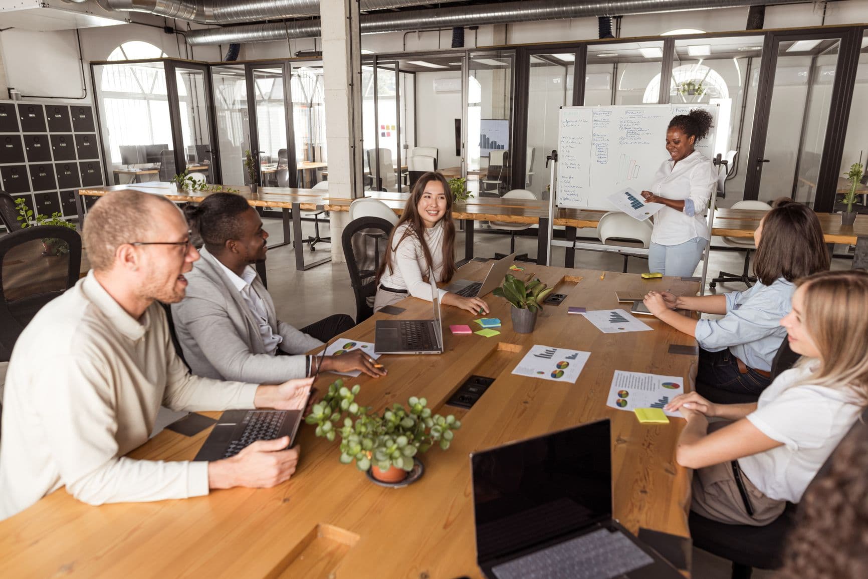 A diverse group of coworkers in a bright office gather around a table and high-five each other, smiling and celebrating teamwork during a collaborative moment.